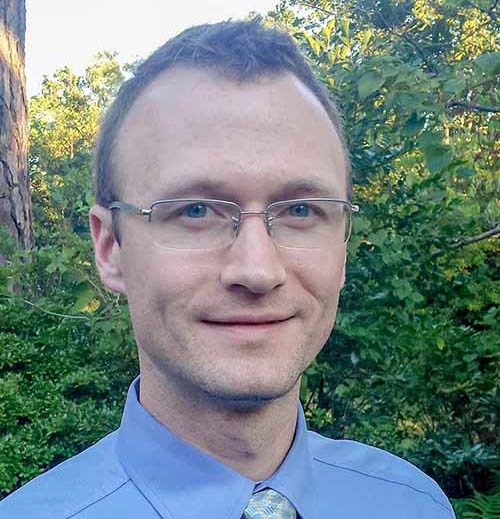A smiling man with glasses and brown hair stands in front of a tree, wearing a blue button-up shirt and tie.