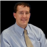A profile of a man with glasses and brown hair wearing a blue button-up shirt and beige tie.