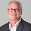 A profile of a smiling man with gray hair and glasses, wearing a black suit jacket and white button-up shirt.