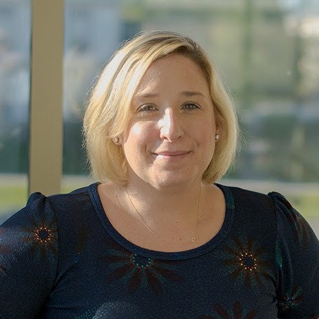 A smiling woman with short blond hair, wearing a dark blue shirt, stands in front of a window.