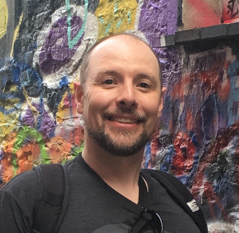 A smiling man with a beard stands in front of a colorful mural wearing a black t-shirt and backpack.