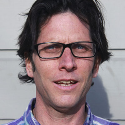 A profile of a man with glasses and dark hair wearing a blue plaid shirt.
