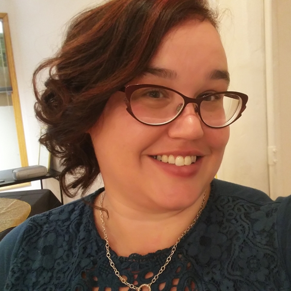 A profile of a smiling woman with dark hair and glasses wearing a silver necklace and teal blouse.