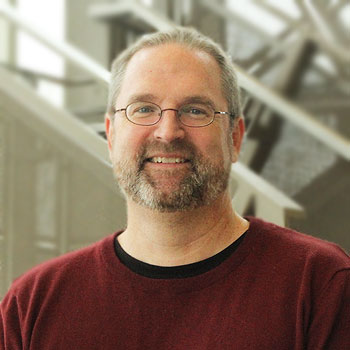 A profile of a smiling man with gray hair and beard, wearing glasses and a red sweater.